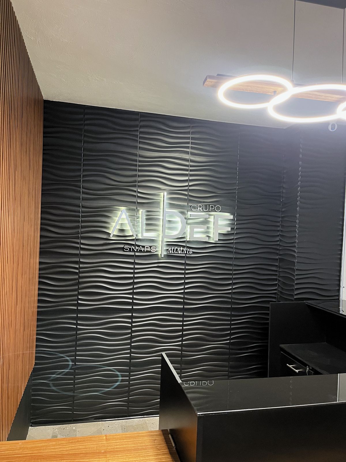 Recepción de Grupo ALDEF con el anuncio luminoso sobre muro de paneles ondulados negros y el mostrador